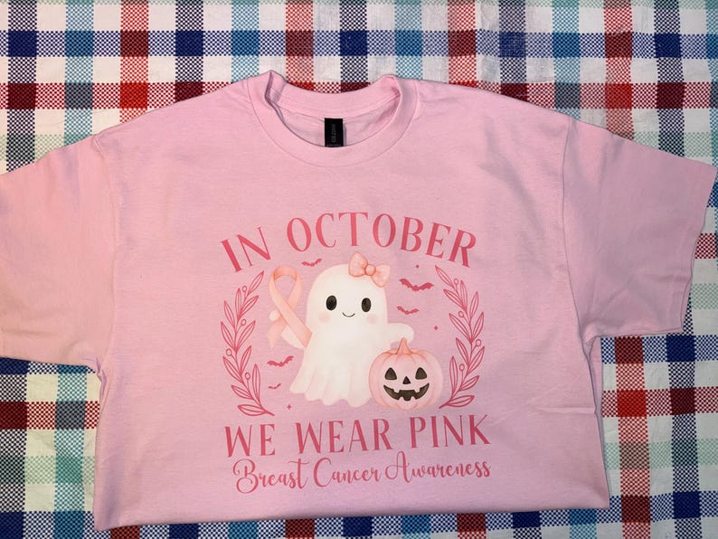 May include: Pink t-shirt with a graphic design featuring a ghost, a pink ribbon, and a jack-o'-lantern. The text reads "In October We Wear Pink Breast Cancer Awareness". The shirt is a crew neck.