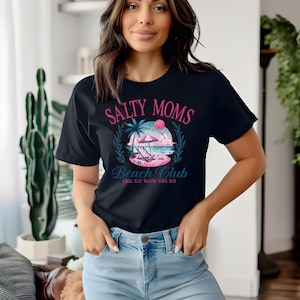 Op de afbeelding: Een zwart T-shirt met een grafisch ontwerp met de tekst "Salty Moms Beach Club Girls Just Wanna Have Sun". De grafiek toont een strandscene met palmbomen, een parasol en een ligstoel.