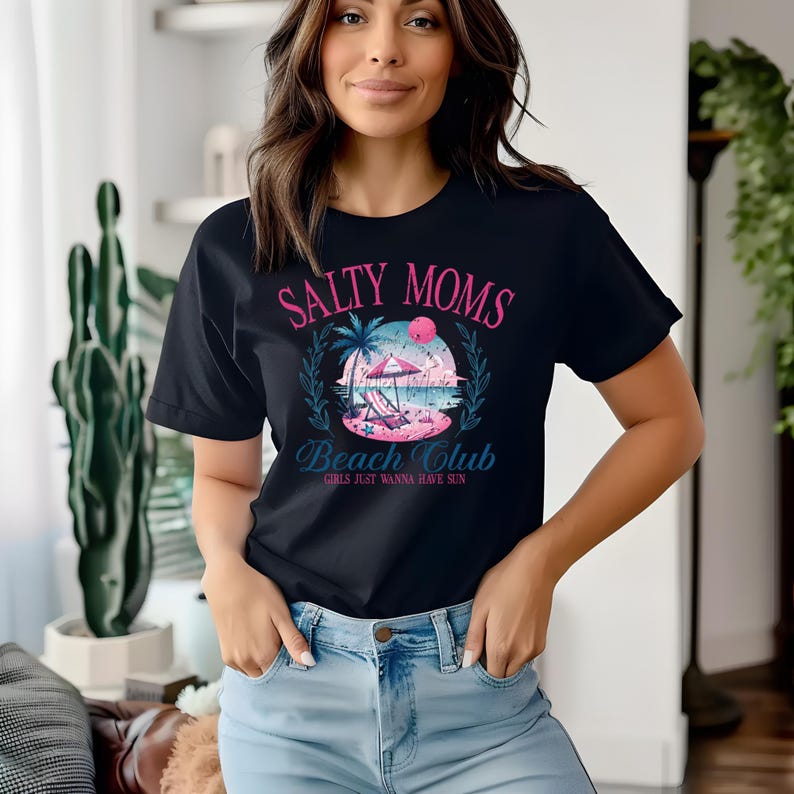 Op de afbeelding: Een zwart T-shirt met een grafisch ontwerp met de tekst "Salty Moms Beach Club Girls Just Wanna Have Sun". De grafiek toont een strandscene met palmbomen, een parasol en een ligstoel.
