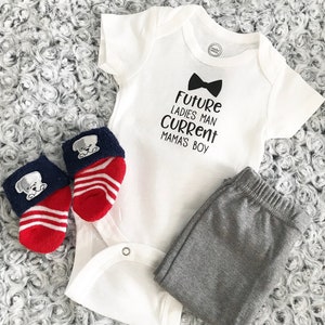 May include: A white baby onesie with the text "Future Ladies Man Current Mama's Boy" and a black bow tie graphic. The outfit includes gray pants and two pairs of socks, one with red and white stripes and the other with a puppy design.