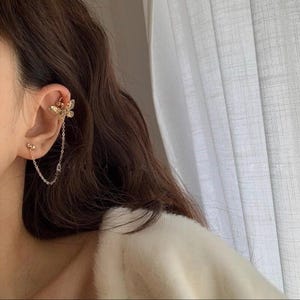 May include: A gold-colored ear cuff featuring a butterfly design, connected by a delicate chain to a stud earring. The butterfly cuff wraps around the upper ear, adding a touch of elegance. The jewelry is shown against a soft, neutral background.