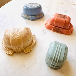 Könnte beinhalten: Vier Schmuckkästchen im Vintage-Stil in Pastellfarben: eine cremefarbene Box mit verzierten Schnörkeln, eine hellblaue Box mit geriffeltem Design, eine korallenfarbene Box mit rechteckigen goldfarbenen Akzenten und eine salbeigrüne Box mit Steppmuster.