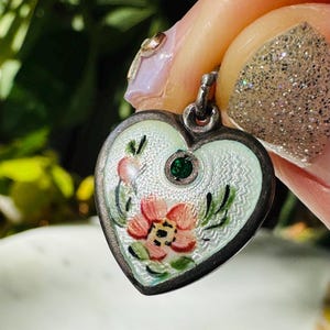 Könnte beinhalten: Herzförmiger Anhänger mit floralem Design. Der Anhänger hat einen silberfarbenen Metallrahmen, einen hellgrünen Hintergrund und eine zentrale Blume mit rosa Blütenblättern und grünen Blättern. Oben ist ein kleiner grüner Edelstein eingesetzt.