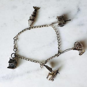 Könnte beinhalten: Ein silbernes Charm-Armband mit verschiedenen detaillierten Charms, darunter ein Fisch, ein Wagen, ein Haus, ein Boot und ein dekoratives Rad. Das Armband hat ein Kettenglied-Design und wird auf einer weißen Marmoroberfläche gezeigt.