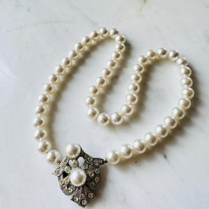 Könnte beinhalten: Eine Vintage-Perlenkette mit einem dekorativen silberfarbenen Anhänger. Der Anhänger hat eine große zentrale Perle, die von kleinen, klaren Kristallen umgeben ist. Die Halskette besteht aus zahlreichen runden, cremeweißen Perlen.