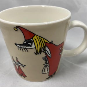 May include: Beige ceramic mug with a white handle. The mug features a whimsical illustration of a character in a red hat and coat with yellow hair. The design is repeated on the side of the mug. The mug has a capacity of approximately 12 ounces.