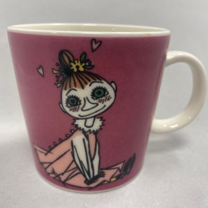 May include: Pink ceramic mug with a white handle featuring a cartoon illustration of a girl with brown hair, green eyes, and a pink dress. The girl is smiling and has hearts around her head.