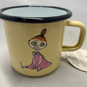 May include: A yellow enamel mug with a black rim and a cartoon illustration of a girl with red hair and a pink dress. The girl is sitting with her legs crossed and has green eyes.