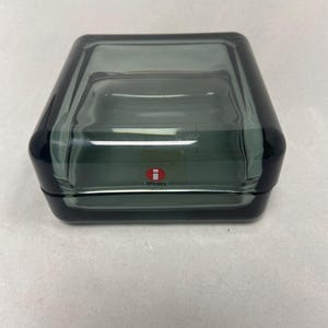 May include: A square, dark grey glass bowl with rounded corners and thick, smooth walls. The center of the front face features a small, square red and white sticker with the text iittala printed in white lowercase letters on the glass surface.
