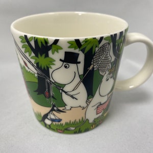 May include: White ceramic mug with a green, black, and white illustration of Moomins fishing in a forest. The mug has a white handle.