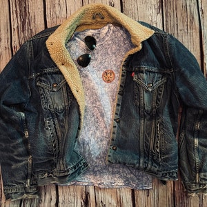 May include: A blue denim jacket with a sherpa lining and a light blue t-shirt underneath. The t-shirt has a faded design and a small circular patch with a design on it.