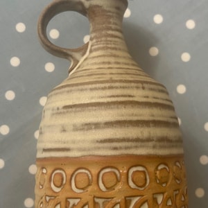 May include: A small, ceramic pitcher with a handle. The pitcher has a light beige and brown striped body, a brown handle, and a decorative band of circles and text near the base. The pitcher is likely used for serving liquids.
