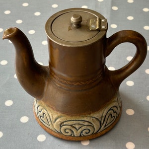 May include: A brown ceramic teapot with a hinged metal lid and a decorative design of hearts and swirls. The teapot has a spout and a handle.