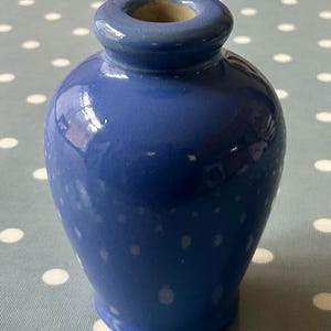 Puede incluir: Un pequeño jarrón de cerámica azul con cuerpo redondeado y abertura ligeramente acampanada. El jarrón tiene un acabado brillante y está decorado con sutiles motas blancas.