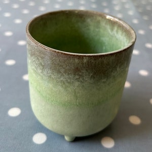 May include: A green ceramic planter with a brown rim and three small feet. The planter has a gradient of green shades, fading from a darker green at the top to a lighter green at the bottom.