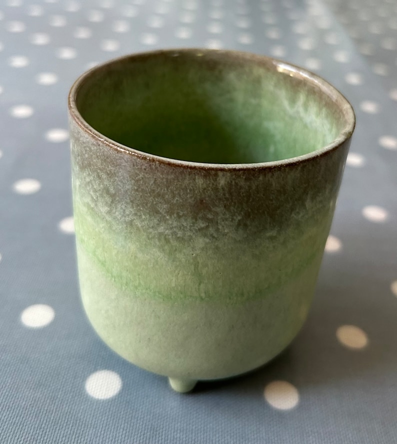 May include: A green ceramic planter with a brown rim and three small feet. The planter has a gradient of green shades, fading from a darker green at the top to a lighter green at the bottom.