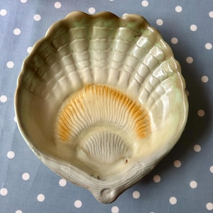 May include: A ceramic seashell-shaped dish with a green and white glaze. The inside of the dish is painted with a yellow and orange design.