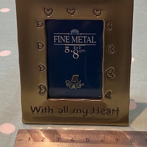 May include: A gold-coloured metal picture frame with a heart design around the border. The frame is 5 inches by 8 inches and has the words "Fine Metal" and "With all my Heart" inscribed on it.