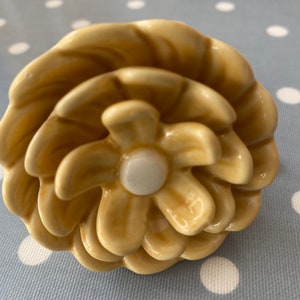 Könnte beinhalten: Ein Keramik-Knopf in Blütenform in blassgelber Farbe mit einem weißen Zentrum. Der Knopf hat ein geschichtetes Design, das an eine Blütenknospe erinnert.
