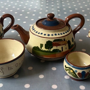 May include: A brown teapot with a white and blue floral design, a white creamer with a blue and brown floral design, a white sugar bowl with a blue and brown floral design, and a white sugar bowl with a blue and brown floral design on a white and grey polka dot tablecloth.