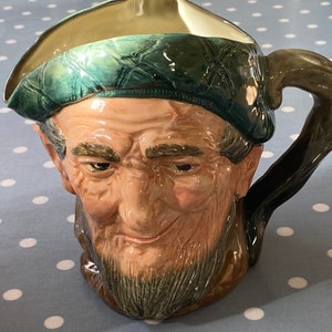 Puede incluir: Una jarra de cerámica con un esmalte verde y marrón. La jarra tiene forma de cabeza de hombre con barba y un sombrero verde.