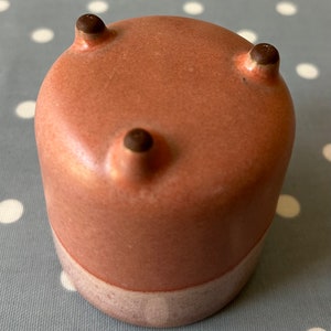 May include: A small, pink ceramic planter with three rounded feet. The planter has a light pink glaze and a white band around the bottom.