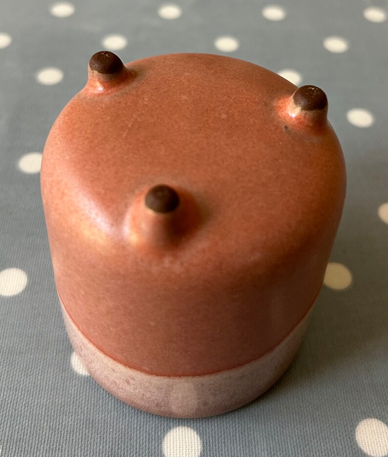 May include: A small, pink ceramic planter with three rounded feet. The planter has a light pink glaze and a white band around the bottom.