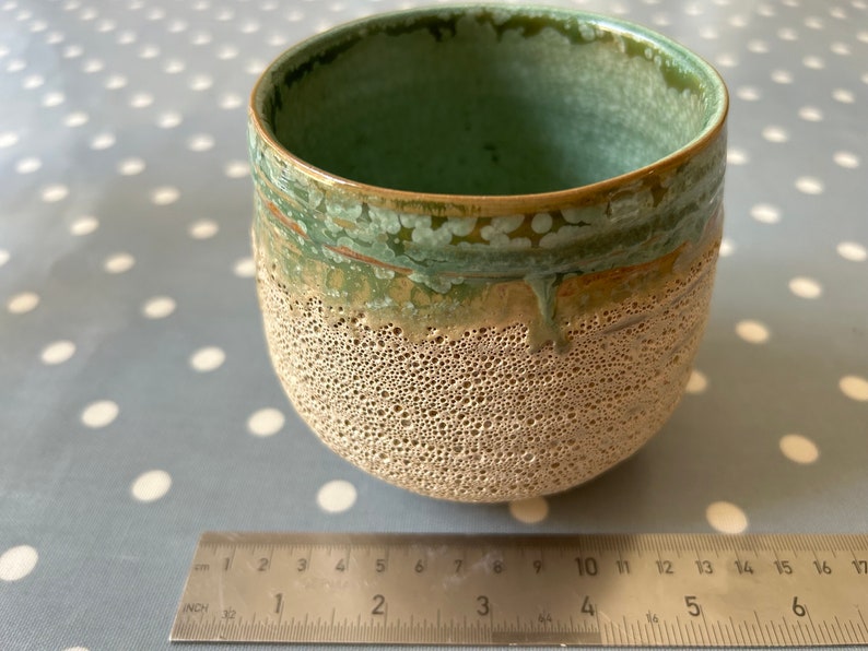 May include: A small, round ceramic planter with a textured, beige exterior and a green glaze on the top rim. The planter is approximately 4 inches in diameter.