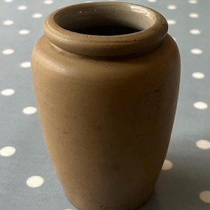 Op de afbeelding: Een kleine, bruine steengoed pot met een rond lichaam en een licht uitlopende lip. De pot heeft een gladde, matte afwerking en is licht beschadigd.