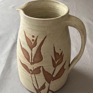 May include: A beige ceramic jug with a handle and spout. The jug is decorated with brown floral designs. The jug is suitable for serving water, juice, or other beverages.