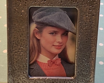 May include: A silver picture frame with a textured surface. The frame holds a photo of a woman wearing a blue beret and a pink sweater.