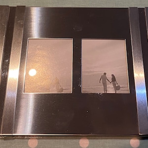 Puede incluir: Un marco de fotos de metal plateado con dos aberturas para fotos. El marco tiene un acabado de metal cepillado y un diseño moderno. El marco está vacío y listo para fotos.