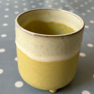 May include: A yellow ceramic planter with a white glaze on the top rim. The planter has three small feet and is perfect for small plants.