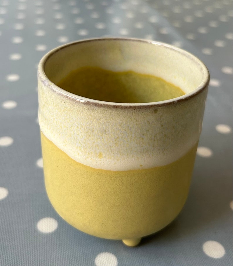May include: A yellow ceramic planter with a white glaze on the top rim. The planter has three small feet and is perfect for small plants.