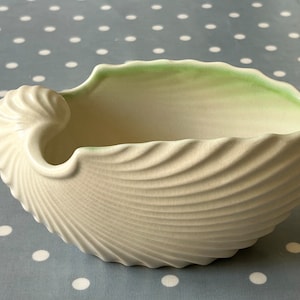May include: A white ceramic seashell-shaped bowl with a green rim. The bowl has a textured, ribbed surface and a slightly open design.