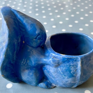 Puede incluir: Un macetero de cerámica azul con forma de conejo con un acabado esmaltado. El macetero tiene una pequeña abertura en la parte superior para plantar.