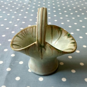 Può includere: Un piccolo cestino in ceramica verde e marrone con un manico. Il cestino ha una finitura smaltata e una superficie leggermente strutturata.