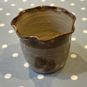 Può includere: Un piccolo vaso in ceramica marrone con un bordo ondulato. Il vaso ha una smaltatura marrone chiaro con una smaltatura marrone più scura intorno al bordo.