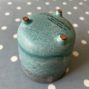 May include: A teal blue ceramic planter with three brown feet. The planter has a speckled glaze and a dark gray base. The bottom of the planter has a white label that reads "by ANTHROPOLOGIE Made in Portugal Fabriqu&eacute; au Portugal".