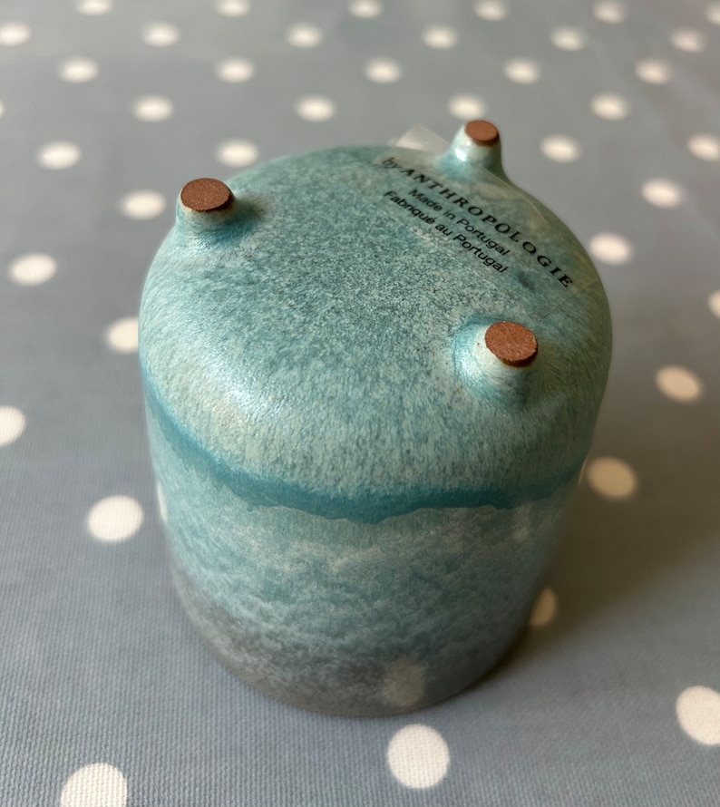 May include: A teal blue ceramic planter with three brown feet. The planter has a speckled glaze and a dark gray base. The bottom of the planter has a white label that reads "by ANTHROPOLOGIE Made in Portugal Fabriqu&eacute; au Portugal".