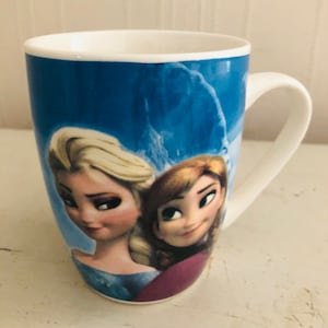 Puede incluir: Una taza de cerámica blanca con fondo azul y una imagen de Elsa y Anna de la película Frozen. La taza tiene un asa blanca.