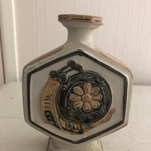 Może przedstawiać: Biały ceramiczny wazon o sześciokątnym kształcie z brązowym i zielonym wzorem ślimaka. Wazon ma brązową krawędź i brązową podstawę.