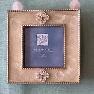 May include: Silver-plated photo frame with a square design. The frame has a cream-coloured, marbled-look border and a smaller square opening. It features decorative crystal accents and the text "SILVERPLATED". The frame is 5x5 cm.