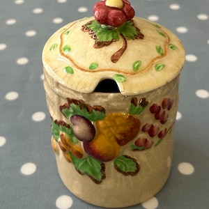 May include: A ceramic jar with a lid, decorated with a floral design and fruit motifs. The jar is cream-coloured with green, brown, yellow, and red accents.
