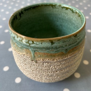 May include: A ceramic bowl with a speckled, light brown exterior and a green glaze on the rim. The bowl has a textured surface and a rounded shape.