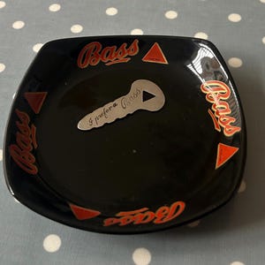 May include: Black ceramic ashtray with red and gold Bass logos and a silver key-shaped bottle opener with the text "I prefer a Bass" on it.