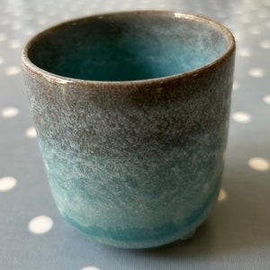 May include: A small, light blue ceramic cup with a brown rim and a speckled glaze. The cup has a rounded bottom and a slightly flared lip.