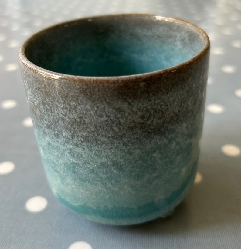 May include: A small, light blue ceramic cup with a brown rim and a speckled glaze. The cup has a rounded bottom and a slightly flared lip.