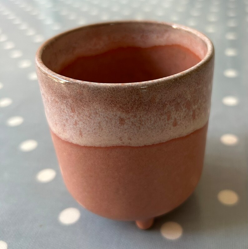 May include: A small, pink ceramic planter with a white glaze on the top rim. The planter has three small feet and is perfect for a small succulent or cactus.