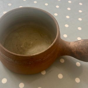 May include: A brown ceramic soup bowl with a handle. The bowl has a round shape with a slightly flared rim and a handle attached to the side. The interior is a lighter colour, and the exterior has a textured finish.